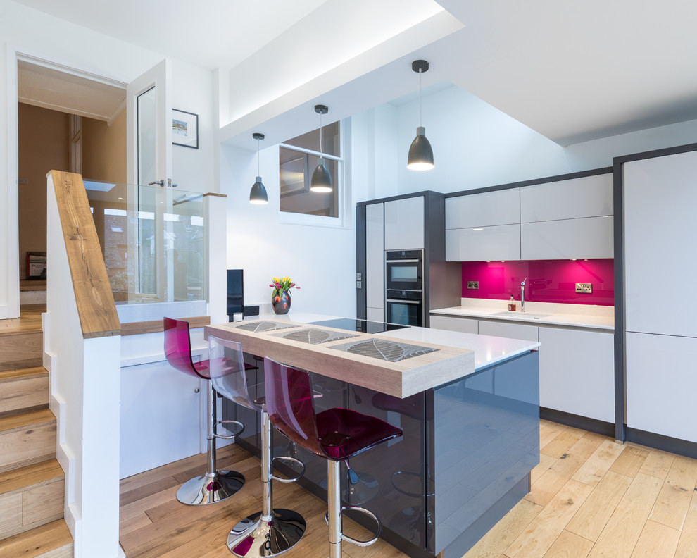 This is an example of a contemporary kitchen in Hampshire with a submerged sink, flat-panel cabinets, white cabinets, pink splashback, black appliances, light hardwood flooring, an island and beige floors.
