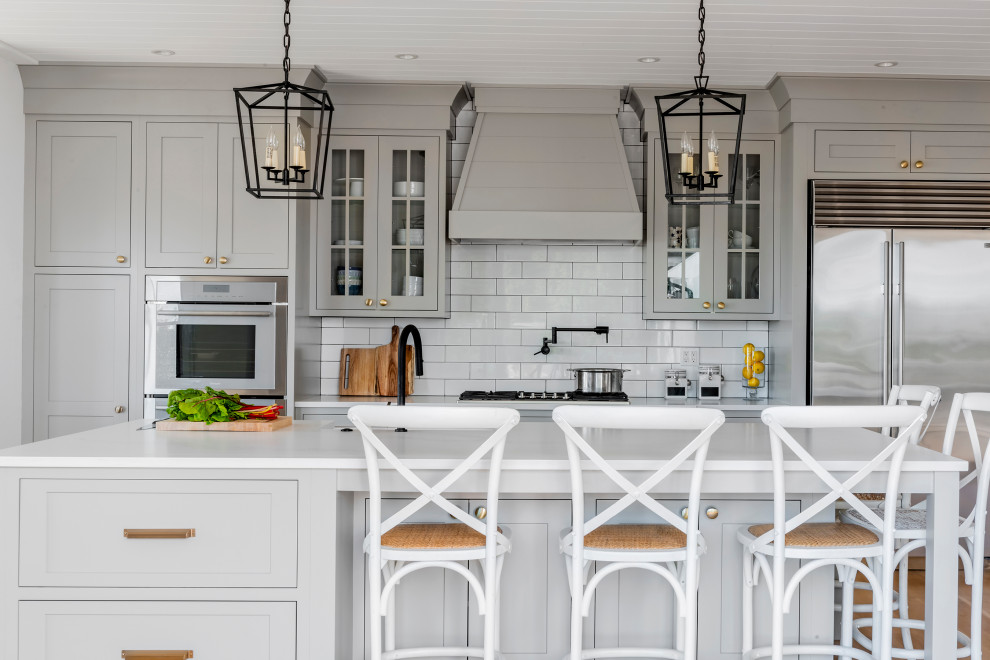 На фото: кухня в стиле неоклассика (современная классика) с стеклянными фасадами, серыми фасадами, белым фартуком, фартуком из плитки кабанчик, техникой из нержавеющей стали, островом и белой столешницей с