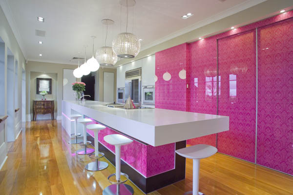 Cette photo montre une grande cuisine parallèle moderne avec un placard à porte plane, un plan de travail en quartz modifié, une crédence rose, une crédence en feuille de verre, un électroménager en acier inoxydable, parquet clair et îlot.