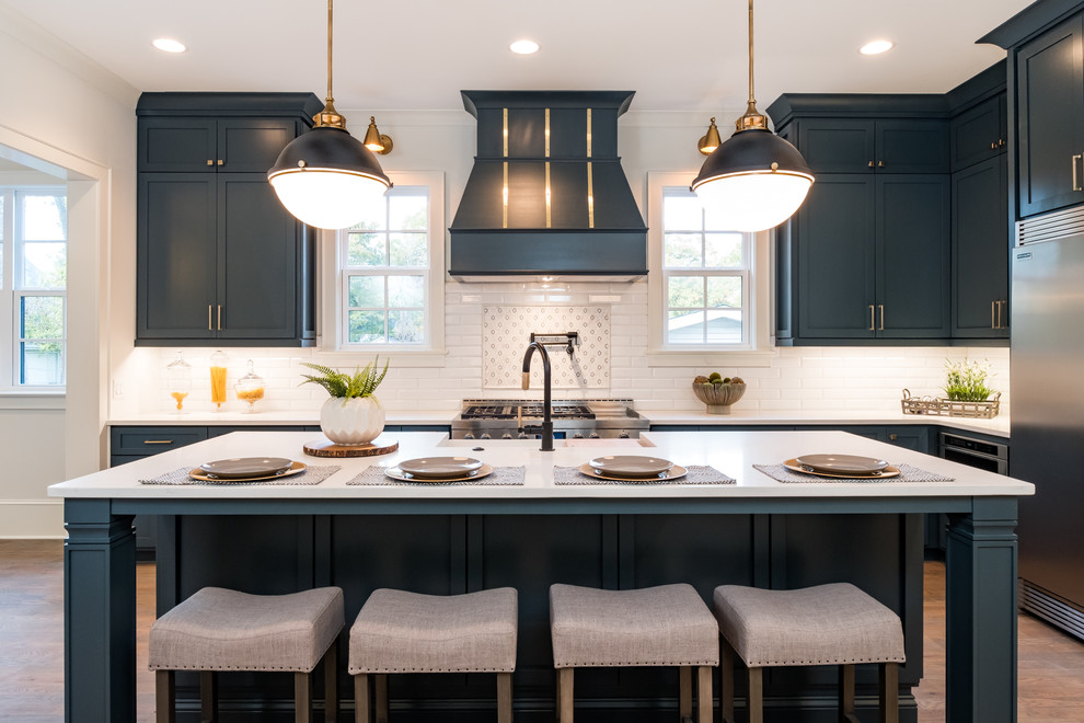 Design ideas for a large country l-shaped kitchen/diner in Charlotte with a belfast sink, blue cabinets, engineered stone countertops, white splashback, glass tiled splashback, stainless steel appliances, medium hardwood flooring, an island, brown floors and white worktops.