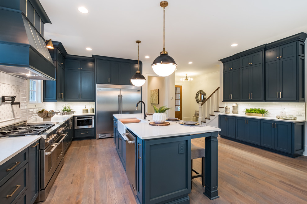 Inspiration for a large country l-shaped kitchen/diner in Charlotte with a belfast sink, blue cabinets, engineered stone countertops, white splashback, glass tiled splashback, stainless steel appliances, medium hardwood flooring, an island, brown floors and white worktops.
