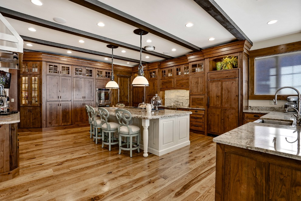 Example of a huge tuscan u-shaped light wood floor and brown floor enclosed kitchen design in Denver with an undermount sink, recessed-panel cabinets, distressed cabinets, granite countertops, beige backsplash, stone slab backsplash, paneled appliances and an island