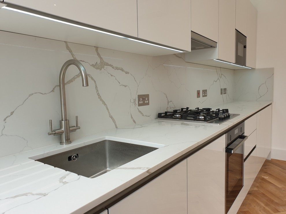 Example of a mid-sized trendy single-wall laminate floor and brown floor open concept kitchen design in London with a drop-in sink, flat-panel cabinets, white cabinets, quartzite countertops, white backsplash, stone slab backsplash, paneled appliances, no island and white countertops