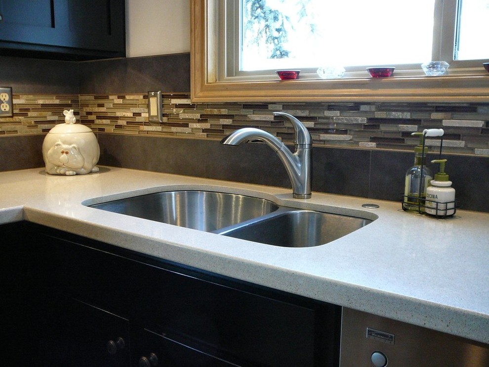 Bild på ett avskilt, litet funkis l-kök, med en undermonterad diskho, släta luckor, svarta skåp, bänkskiva i kvarts, grått stänkskydd, stänkskydd i mosaik och rostfria vitvaror