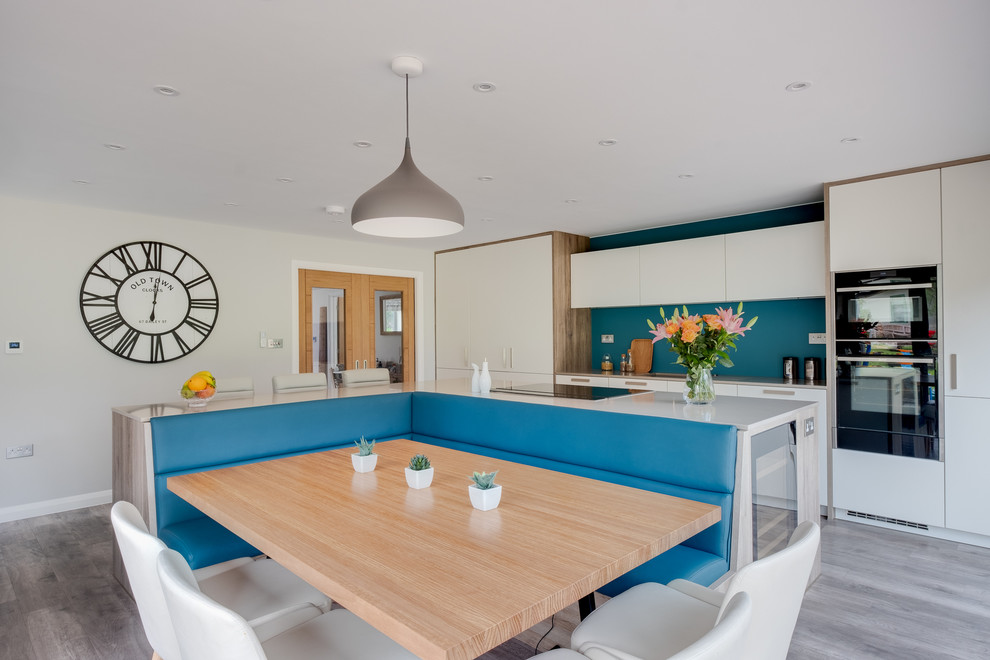 Photo of a large scandinavian kitchen/diner in Gloucestershire with a belfast sink, flat-panel cabinets, white cabinets, composite countertops, blue splashback, glass sheet splashback, black appliances, porcelain flooring, an island, brown floors and white worktops.