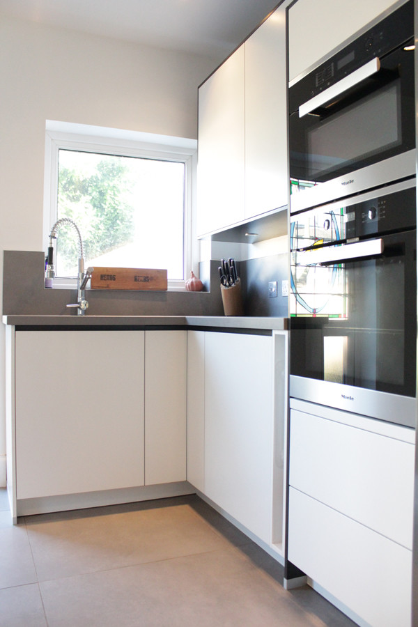 This is an example of a small modern l-shaped kitchen/diner in London with a built-in sink, flat-panel cabinets, white cabinets, granite worktops, grey splashback, porcelain splashback, black appliances, porcelain flooring and an island.