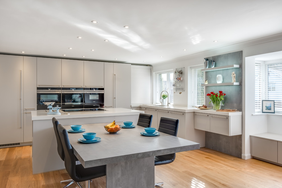 Open concept kitchen - large contemporary l-shaped light wood floor and beige floor open concept kitchen idea in Gloucestershire with an undermount sink, flat-panel cabinets, gray cabinets, black appliances, solid surface countertops and an island