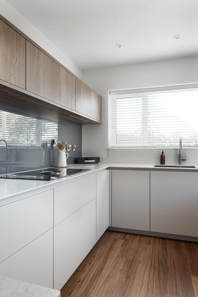 Modern inredning av ett mellanstort vit vitt kök, med en undermonterad diskho, släta luckor, grå skåp, bänkskiva i kvartsit, grått stänkskydd, glaspanel som stänkskydd, svarta vitvaror, mellanmörkt trägolv, en halv köksö och brunt golv