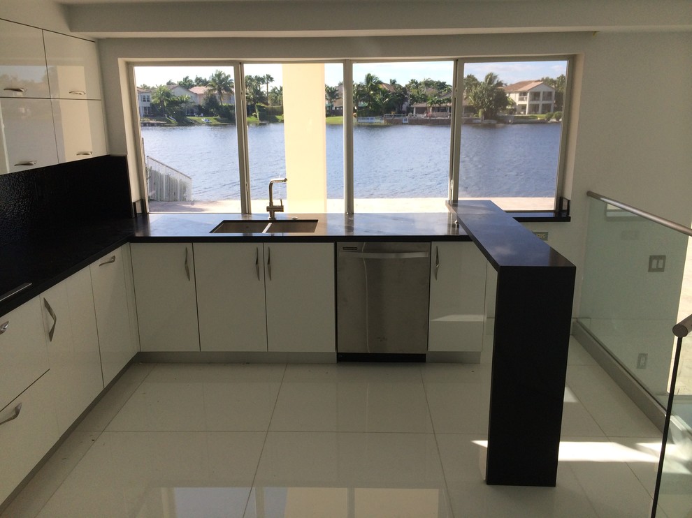 Inspiration for a large contemporary galley open plan kitchen in Miami with a double-bowl sink, flat-panel cabinets, white cabinets, engineered stone countertops, black splashback, stone slab splashback, stainless steel appliances, marble flooring and a breakfast bar.