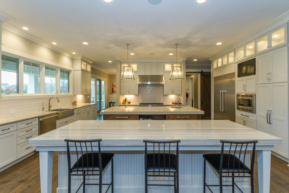 Huge trendy u-shaped dark wood floor and brown floor enclosed kitchen photo in Indianapolis with a farmhouse sink, shaker cabinets, white cabinets, marble countertops, white backsplash, subway tile backsplash, stainless steel appliances and two islands