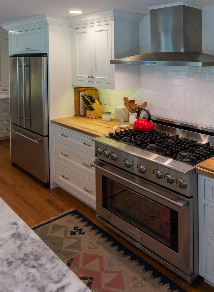 高級な広いトランジショナルスタイルのおしゃれなキッチン (アンダーカウンターシンク、フラットパネル扉のキャビネット、白いキャビネット、木材カウンター、白いキッチンパネル、セラミックタイルのキッチンパネル、シルバーの調理設備、無垢フローリング、茶色い床、茶色いキッチンカウンター) の写真