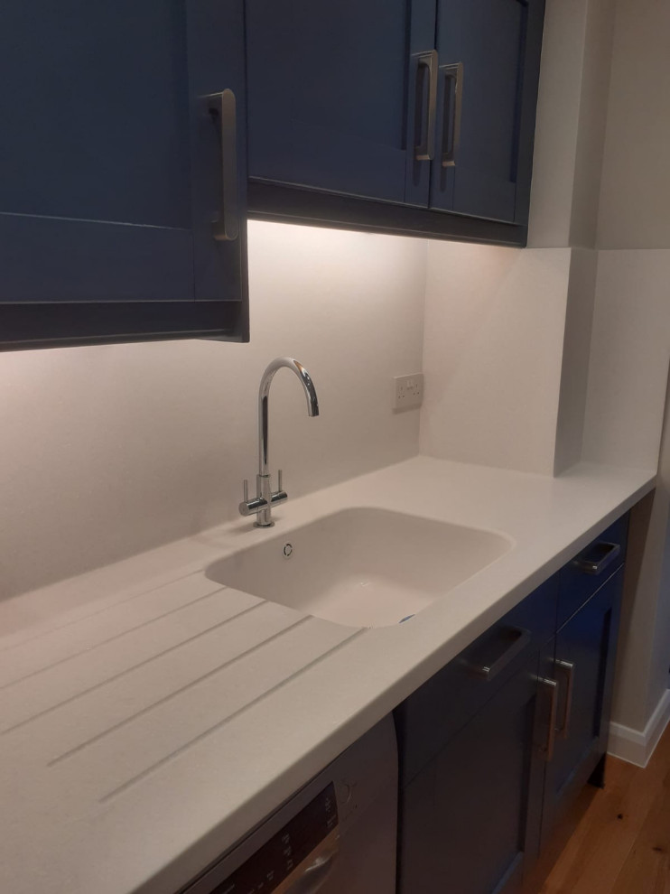 Cette image montre une cuisine américaine design en L de taille moyenne avec un placard à porte shaker, des portes de placard bleues, une crédence blanche et un plan de travail blanc.