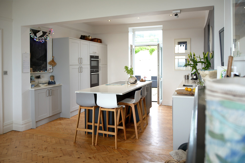Große, Zweizeilige Klassische Wohnküche mit Unterbauwaschbecken, Schrankfronten im Shaker-Stil, grauen Schränken, Mineralwerkstoff-Arbeitsplatte, Küchengeräten aus Edelstahl, braunem Holzboden, Kücheninsel, weißer Arbeitsplatte, Küchenrückwand in Weiß und braunem Boden in Cornwall
