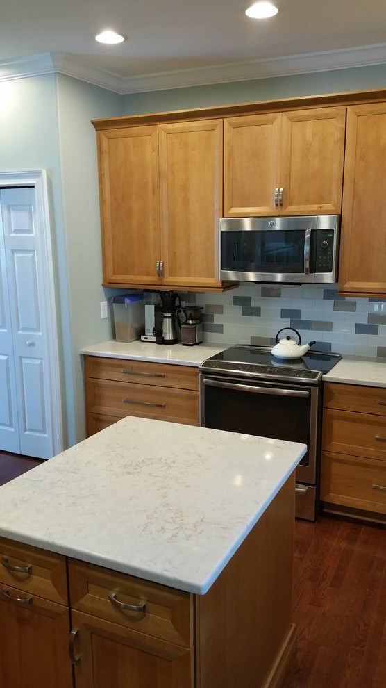 Foto på ett stort vit kök, med luckor med infälld panel, skåp i ljust trä, bänkskiva i kvarts, grått stänkskydd, stänkskydd i glaskakel, rostfria vitvaror, mellanmörkt trägolv, en köksö och brunt golv