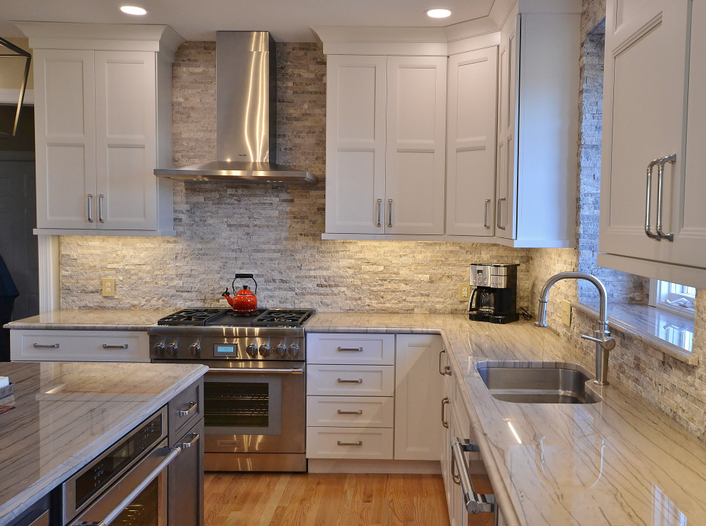 This is an example of a large l-shaped kitchen/diner in Philadelphia with a submerged sink, raised-panel cabinets, white cabinets, quartz worktops, grey splashback, stone tiled splashback, stainless steel appliances, medium hardwood flooring, an island, beige floors and white worktops.