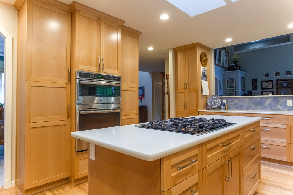 Example of a mid-sized classic l-shaped light wood floor and beige floor enclosed kitchen design with shaker cabinets, light wood cabinets, quartz countertops, blue backsplash, stone tile backsplash, stainless steel appliances, an island and beige countertops