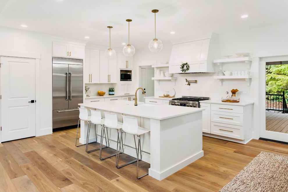 Photo of a medium sized classic u-shaped kitchen/diner in Los Angeles with a belfast sink, shaker cabinets, white cabinets, white splashback, stainless steel appliances, medium hardwood flooring, an island, brown floors, white worktops, engineered stone countertops and metro tiled splashback.