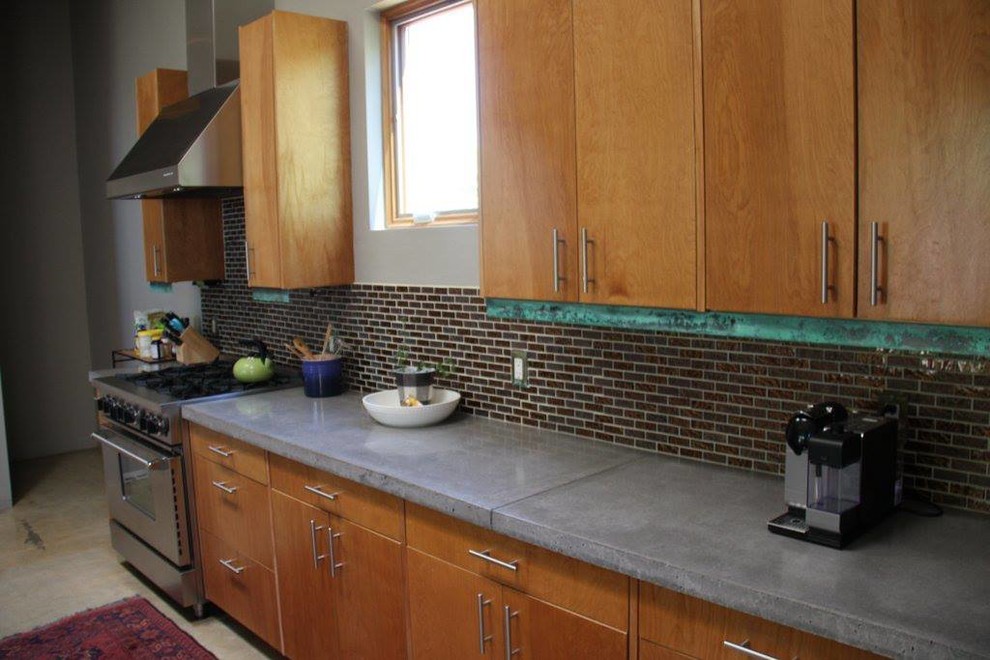Inredning av ett modernt stort kök och matrum, med en undermonterad diskho, skåp i mellenmörkt trä, bänkskiva i betong, stänkskydd med metallisk yta, rostfria vitvaror, betonggolv och en köksö