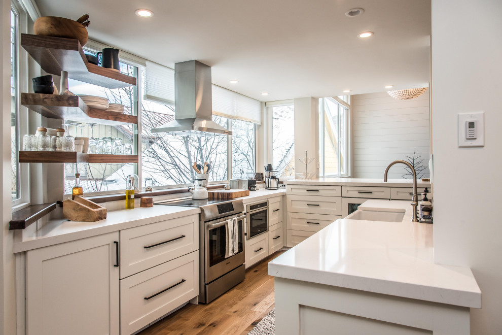 Foto på ett funkis vit l-kök, med en rustik diskho, skåp i shakerstil, vita skåp, rostfria vitvaror, mellanmörkt trägolv, en köksö och brunt golv