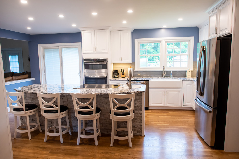 Inredning av ett klassiskt mellanstort blå blått kök, med en rustik diskho, luckor med upphöjd panel, vita skåp, bänkskiva i kvarts, grått stänkskydd, stänkskydd i glaskakel, rostfria vitvaror, mellanmörkt trägolv, en köksö och brunt golv