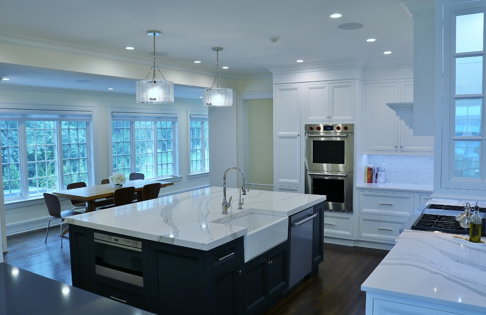 Inredning av ett klassiskt stort vit vitt kök, med en rustik diskho, luckor med infälld panel, vita skåp, bänkskiva i kvarts, stänkskydd i glaskakel, rostfria vitvaror, mellanmörkt trägolv, en köksö och brunt golv