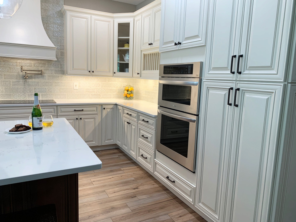 Example of a farmhouse laminate floor, brown floor and tray ceiling eat-in kitchen design in Orlando with a farmhouse sink, raised-panel cabinets, white cabinets, quartz countertops, blue backsplash, ceramic backsplash, stainless steel appliances, an island and white countertops