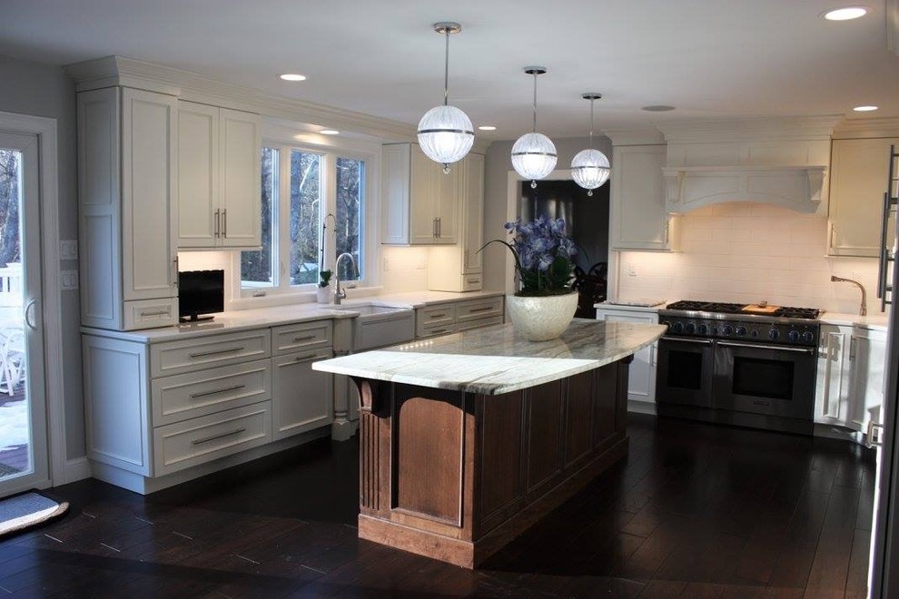 This is an example of a large classic kitchen in Boston with a belfast sink, recessed-panel cabinets, white cabinets, white splashback, metro tiled splashback, stainless steel appliances, medium hardwood flooring, an island and brown floors.