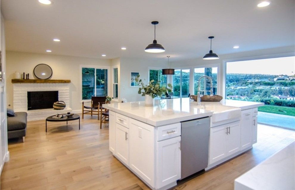 This is an example of a medium sized modern l-shaped open plan kitchen in Los Angeles with a belfast sink, shaker cabinets, white cabinets, engineered stone countertops, white splashback, metro tiled splashback, stainless steel appliances, light hardwood flooring, an island, beige floors and white worktops.