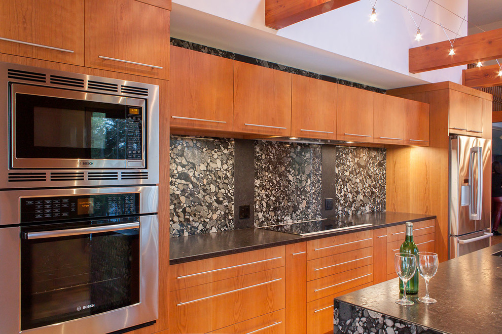 Cette image montre une cuisine américaine linéaire design en bois brun avec un évier de ferme, un placard à porte plane, un plan de travail en granite, une crédence noire et un électroménager en acier inoxydable.