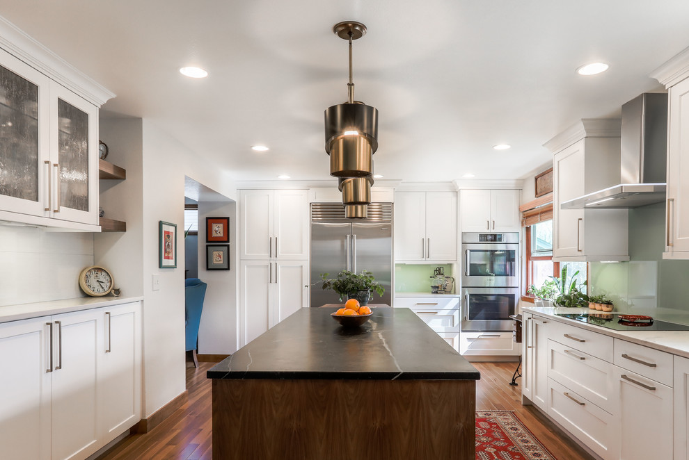 Example of a transitional u-shaped medium tone wood floor and brown floor kitchen design in Denver with shaker cabinets, white cabinets, green backsplash, glass sheet backsplash, stainless steel appliances, an island and beige countertops