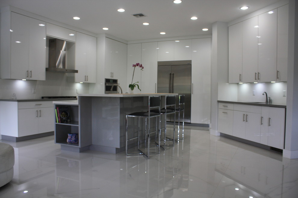 Photo of a large contemporary l-shaped enclosed kitchen in Miami with flat-panel cabinets, white cabinets, engineered stone countertops, white splashback, stone slab splashback, stainless steel appliances, an island, white floors, a submerged sink, marble flooring and white worktops.