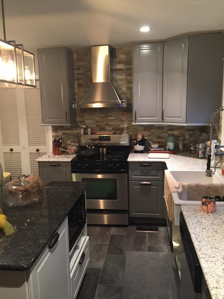 Photo of a large contemporary l-shaped kitchen/diner in Indianapolis with a belfast sink, raised-panel cabinets, grey cabinets, granite worktops, beige splashback, brick splashback, stainless steel appliances, cement flooring and an island.