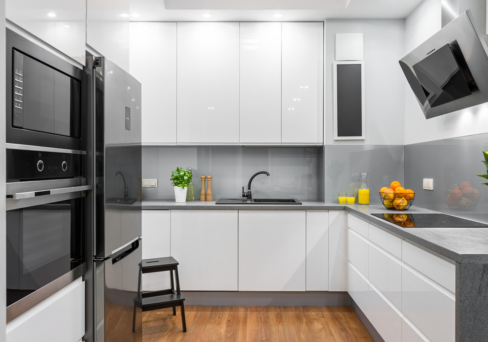 This is an example of a contemporary grey and white l-shaped kitchen in Los Angeles with a built-in sink, flat-panel cabinets, white cabinets, stainless steel appliances, medium hardwood flooring and brown floors.
