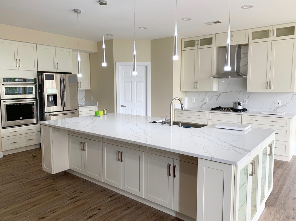 Large modern l-shaped kitchen/diner in DC Metro with a double-bowl sink, shaker cabinets, white cabinets, engineered stone countertops, white splashback, stone slab splashback, stainless steel appliances, vinyl flooring, an island, brown floors and white worktops.
