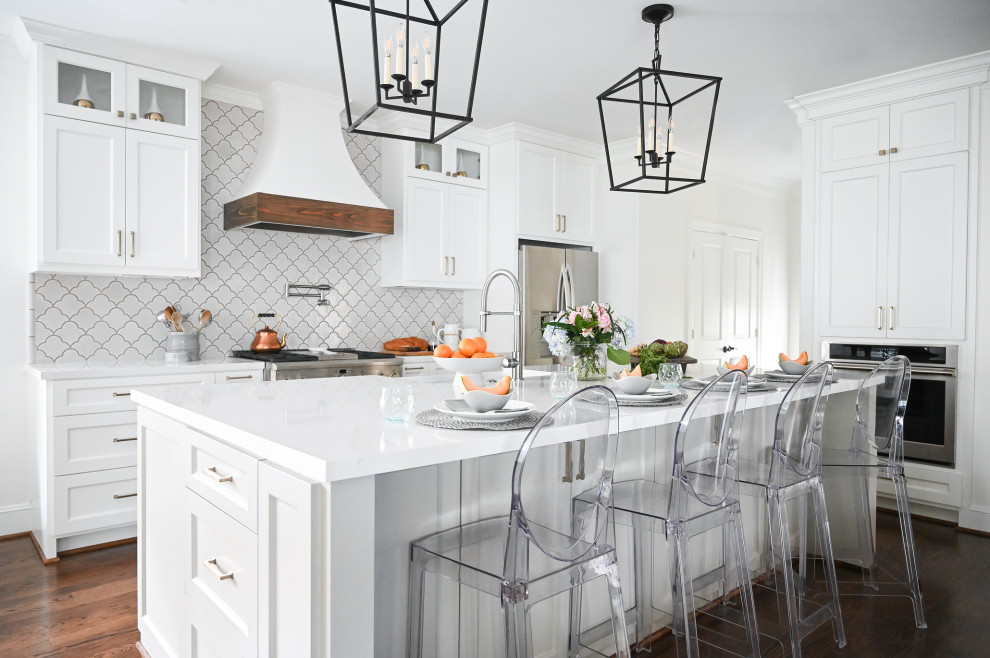 Example of a transitional u-shaped medium tone wood floor and brown floor kitchen design in Houston with a farmhouse sink, shaker cabinets, white cabinets, white backsplash, stainless steel appliances, an island and white countertops