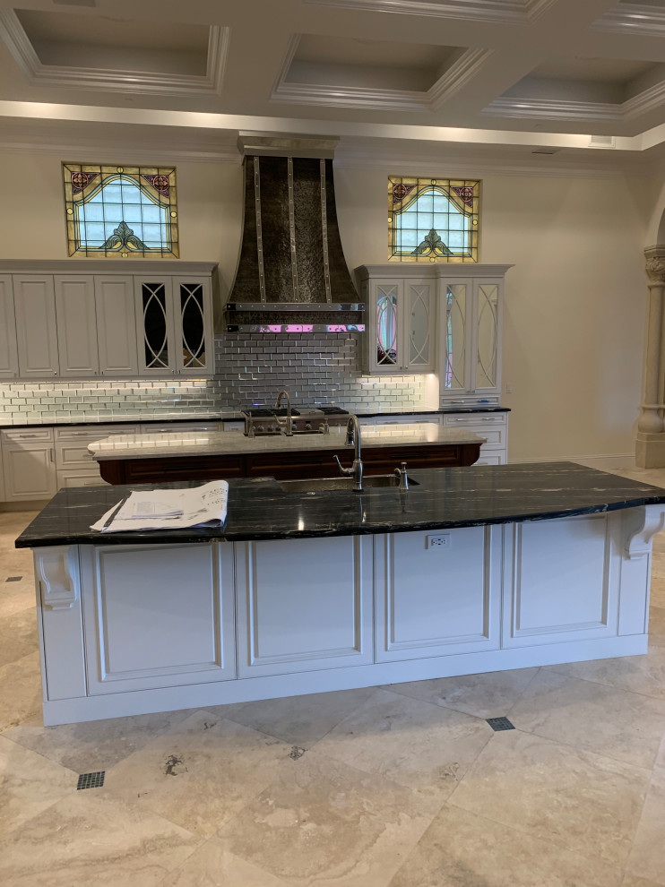 This is an example of a large traditional l-shaped kitchen in Miami with a belfast sink, raised-panel cabinets, white cabinets, granite worktops, metallic splashback, mirror splashback, integrated appliances, marble flooring, an island, beige floors, black worktops and a coffered ceiling.