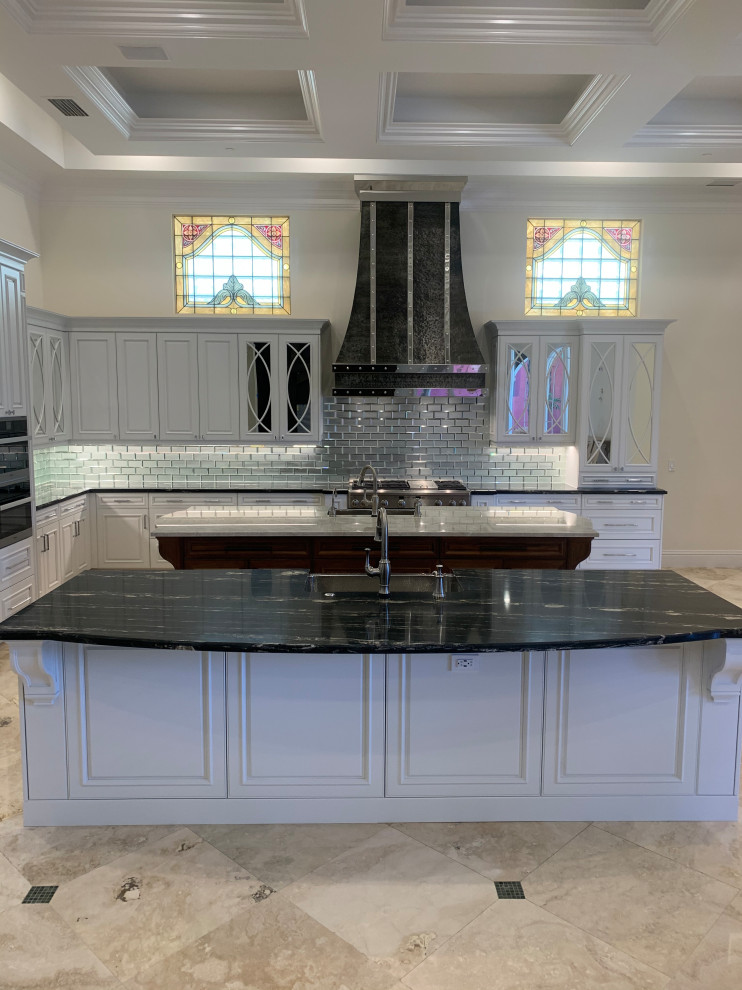 Large classic l-shaped kitchen in Miami with a belfast sink, raised-panel cabinets, white cabinets, granite worktops, metallic splashback, mirror splashback, integrated appliances, marble flooring, an island, beige floors, black worktops and a coffered ceiling.