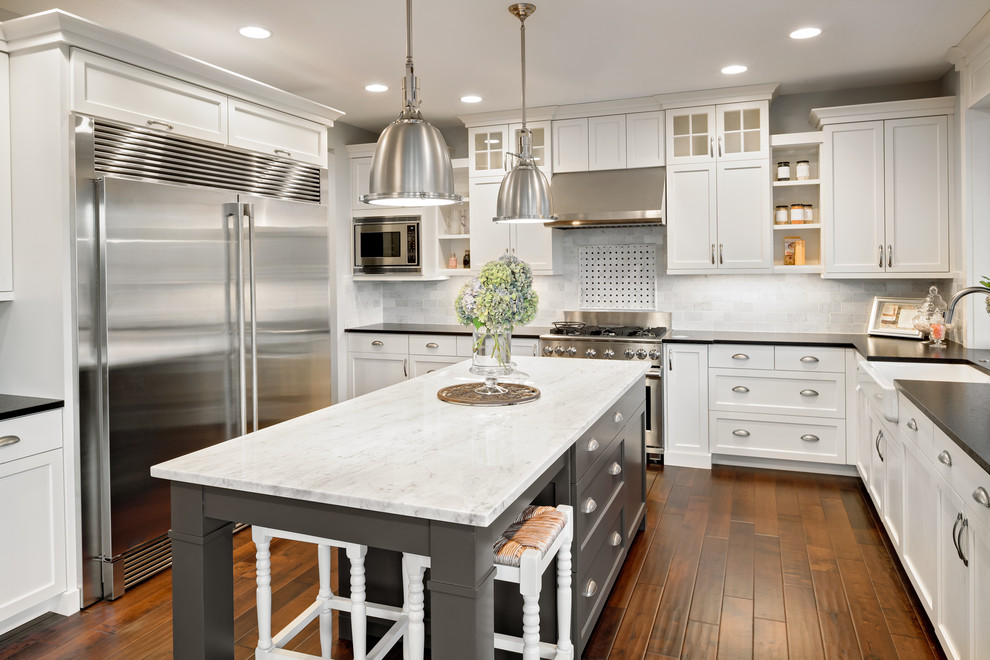 Inspiration for a large transitional u-shaped medium tone wood floor and brown floor eat-in kitchen remodel in Other with a farmhouse sink, shaker cabinets, white cabinets, marble countertops, gray backsplash, stone tile backsplash, stainless steel appliances and an island