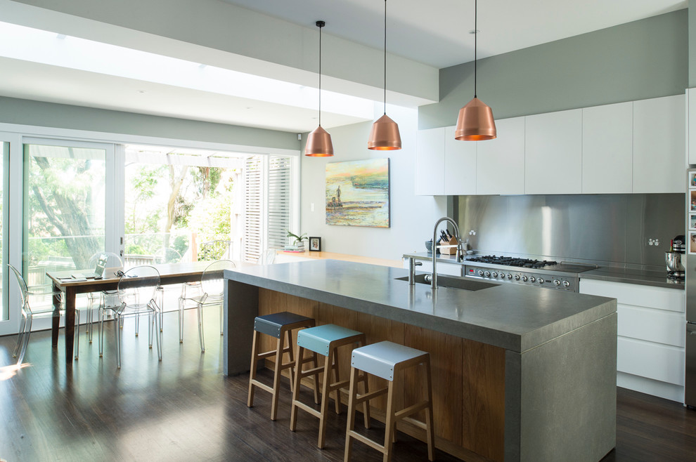 Modern inredning av ett kök, med en undermonterad diskho, vita skåp, bänkskiva i betong, stänkskydd med metallisk yta, rostfria vitvaror, mörkt trägolv, en köksö och släta luckor