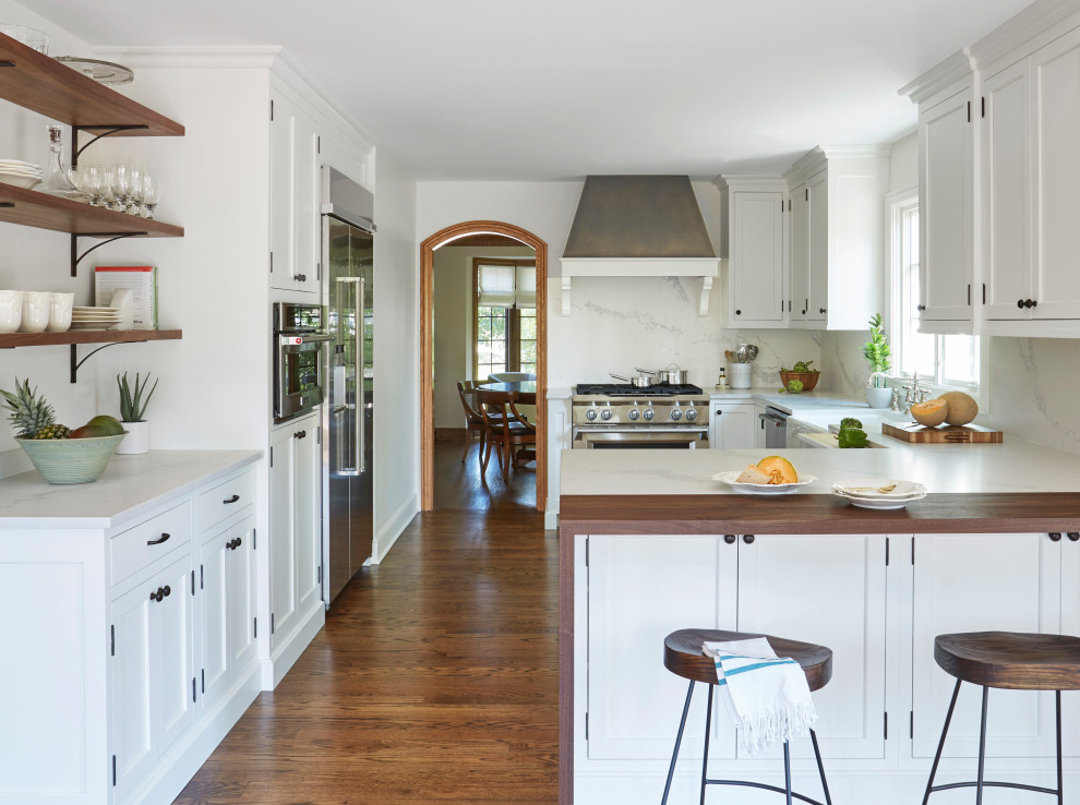 Example of a transitional u-shaped dark wood floor and brown floor kitchen design in Chicago with a farmhouse sink, recessed-panel cabinets, white cabinets, white backsplash, stone slab backsplash, stainless steel appliances, a peninsula and white countertops