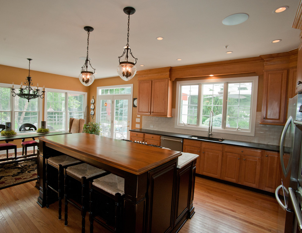 Example of a large classic l-shaped light wood floor and brown floor eat-in kitchen design in Philadelphia with an undermount sink, raised-panel cabinets, beige backsplash, subway tile backsplash, stainless steel appliances, an island, medium tone wood cabinets, solid surface countertops and black countertops