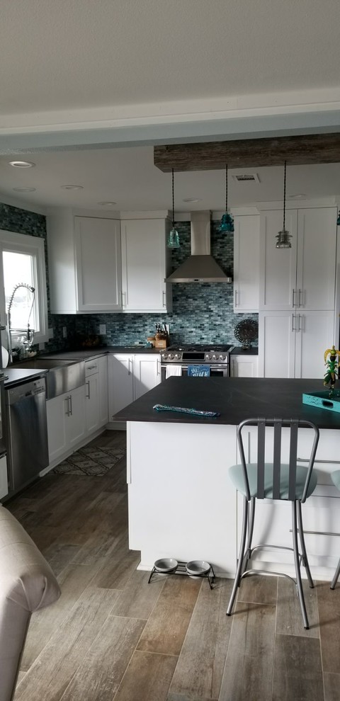 Photo of a large rustic u-shaped kitchen/diner in Houston with a belfast sink, shaker cabinets, white cabinets, engineered stone countertops, blue splashback, glass tiled splashback, stainless steel appliances, porcelain flooring, an island, beige floors and black worktops.