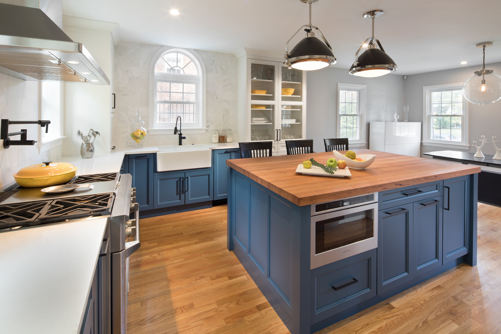 This is an example of a large traditional l-shaped kitchen/diner in DC Metro with a belfast sink, blue cabinets, light hardwood flooring, an island, white splashback, stainless steel appliances, shaker cabinets and engineered stone countertops.