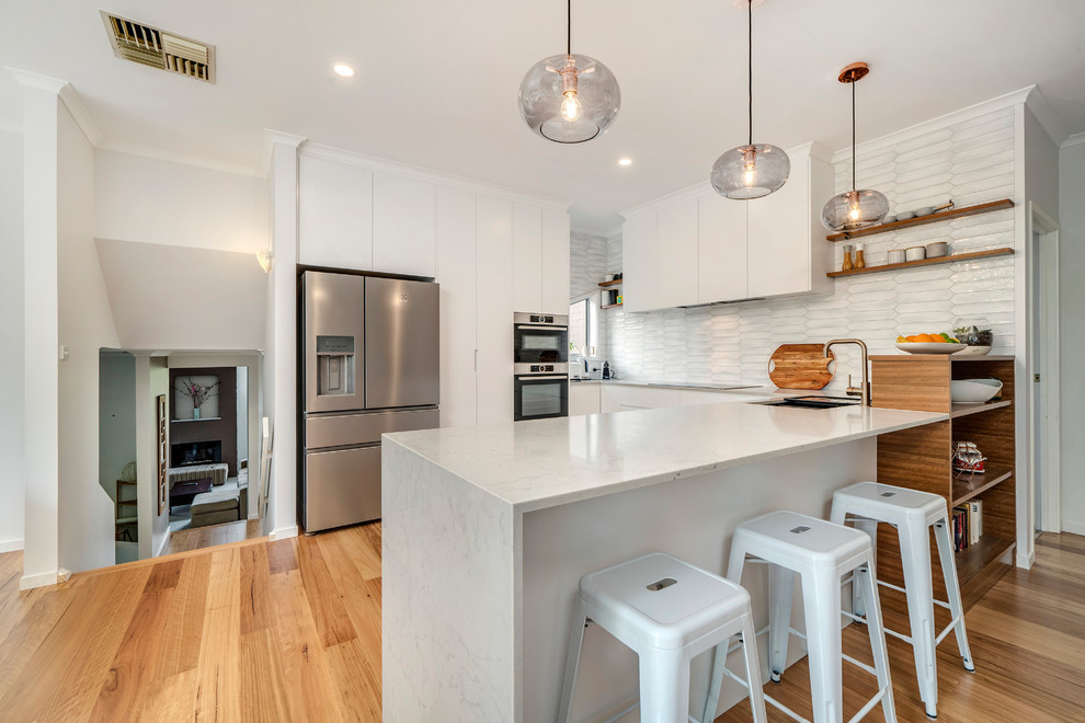 This is an example of a medium sized contemporary u-shaped open plan kitchen in Canberra - Queanbeyan with a submerged sink, white cabinets, engineered stone countertops, grey splashback, metro tiled splashback, stainless steel appliances, light hardwood flooring and white worktops.