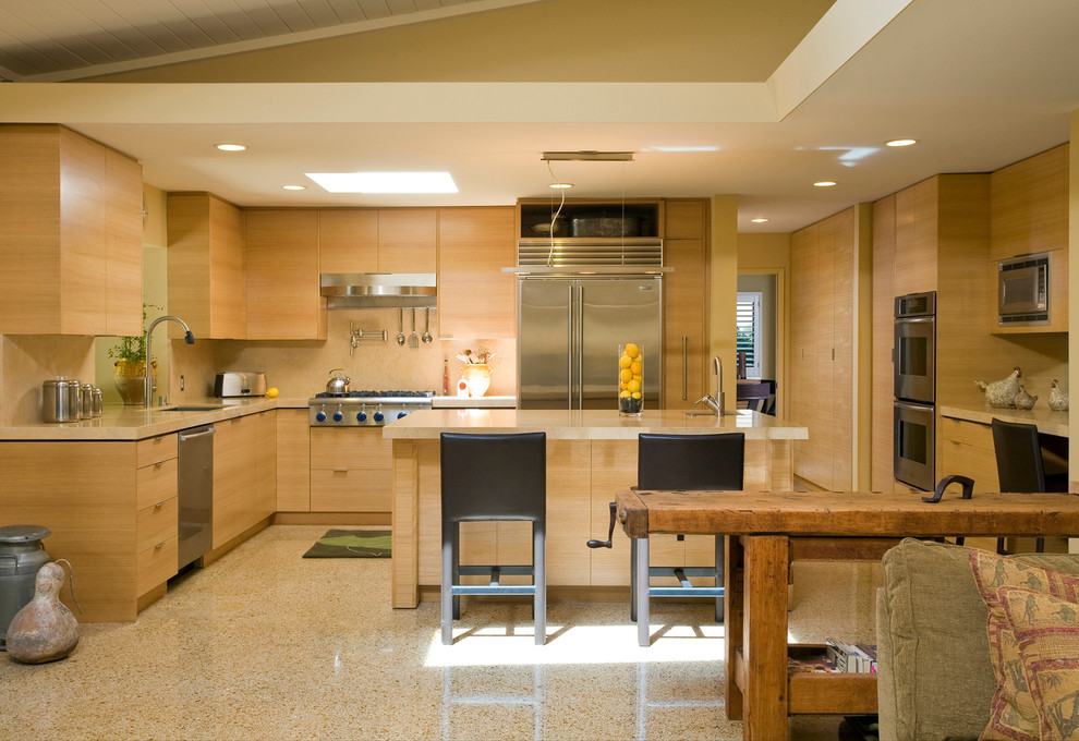 На фото: угловая кухня в современном стиле с плоскими фасадами, светлыми деревянными фасадами, бежевым фартуком и техникой из нержавеющей стали с