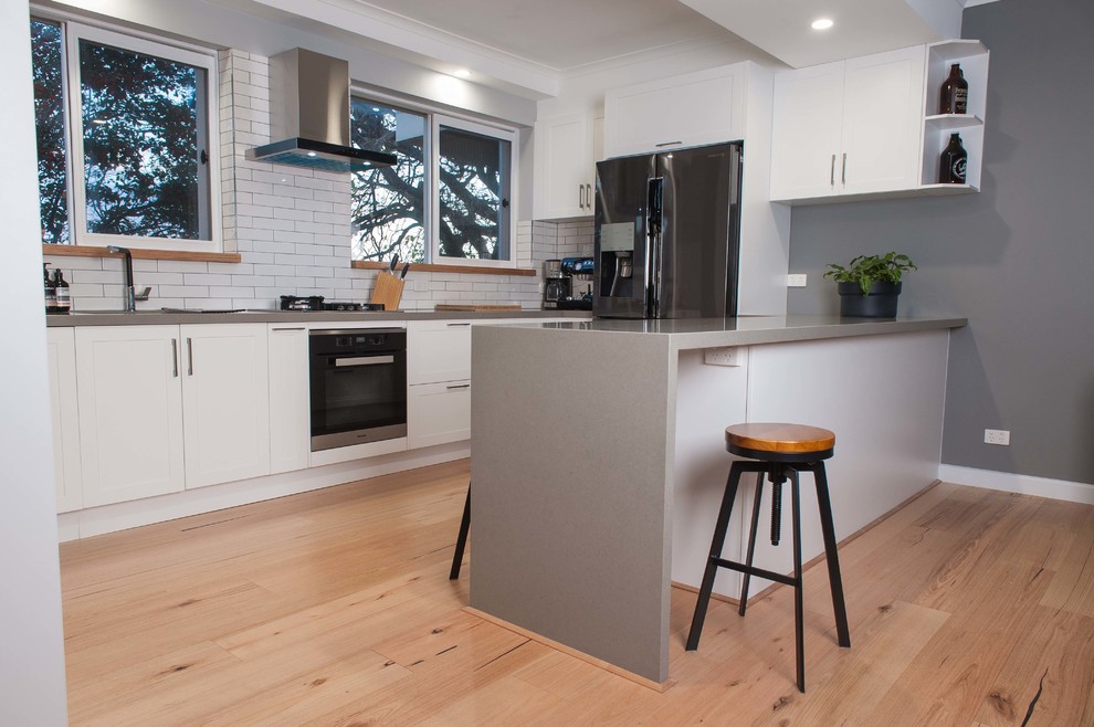 Medium sized scandinavian u-shaped open plan kitchen in Melbourne with a submerged sink, shaker cabinets, white cabinets, composite countertops, white splashback, metro tiled splashback, stainless steel appliances, light hardwood flooring and an island.