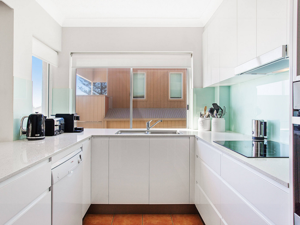 На фото: маленькая п-образная кухня в стиле модернизм с врезной мойкой, плоскими фасадами, белыми фасадами, фартуком из стекла, оранжевым полом и серой столешницей для на участке и в саду с