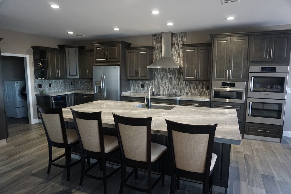 Modern inredning av ett stort grå grått kök, med en undermonterad diskho, luckor med upphöjd panel, grå skåp, granitbänkskiva, grått stänkskydd, stänkskydd i mosaik, rostfria vitvaror, klinkergolv i keramik, en köksö och grått golv