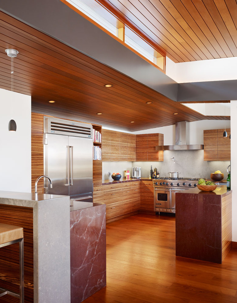 Bild på ett stort tropiskt kök, med rostfria vitvaror, släta luckor, skåp i mörkt trä, marmorbänkskiva, grått stänkskydd, mörkt trägolv, en halv köksö och brunt golv