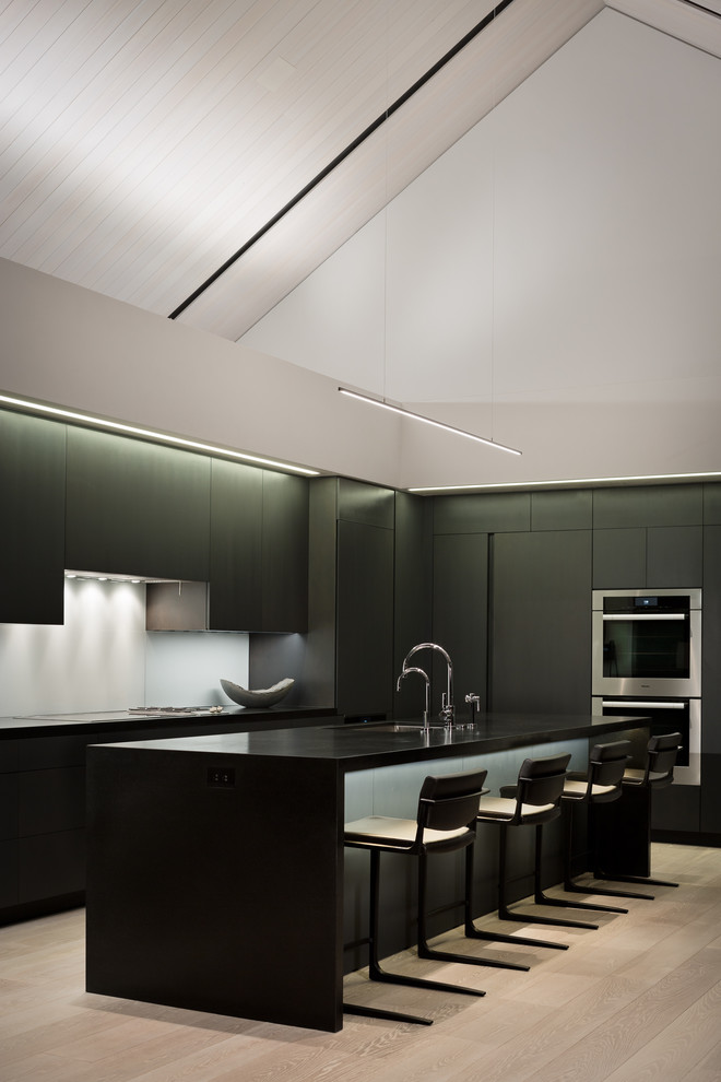 Example of a huge minimalist l-shaped light wood floor and white floor open concept kitchen design in Denver with an undermount sink, flat-panel cabinets, black cabinets, concrete countertops, gray backsplash, glass sheet backsplash, stainless steel appliances, an island and black countertops